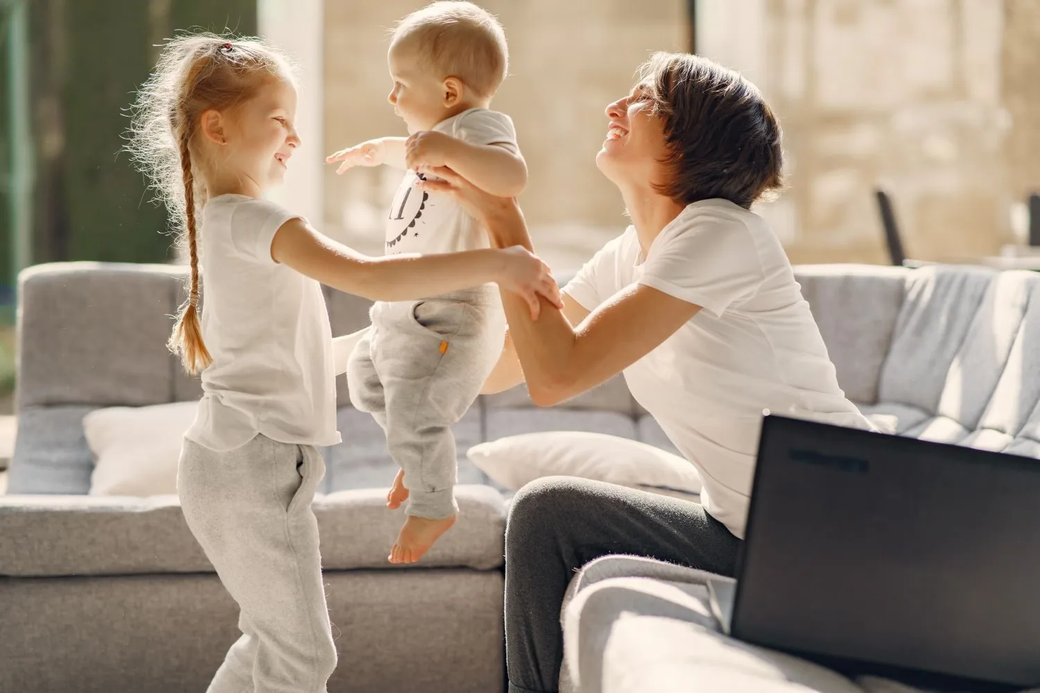 A parent laughing while lifting a small child, with an older child beside them at home.