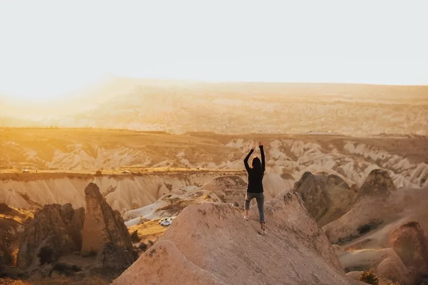 A person standing on a hilltop with both arms raised as the sun rises.